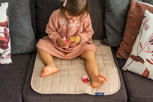 Child sitting on a beige mat on a couch with toys around. Brolly sheets Waterproof Chair Pad.