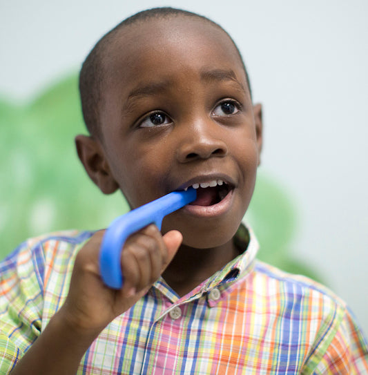 Child chewing on ark grabber sensory tool against a blurred green and white background