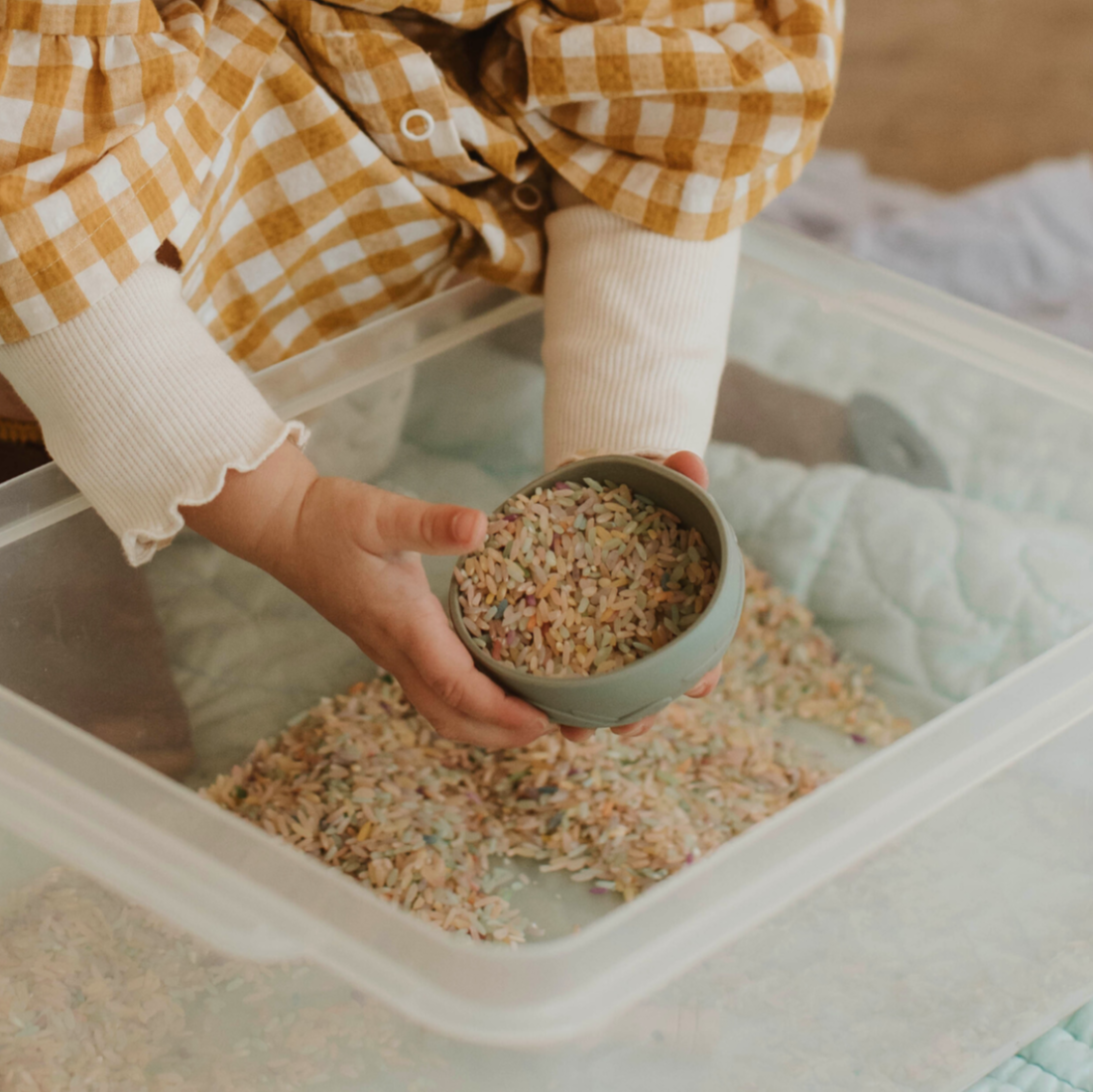 Child engaging in hands-on sensory play using wooden scoop and rainbow rice in sensory bin – supports emotional regulation and NDIS-eligible therapy goals