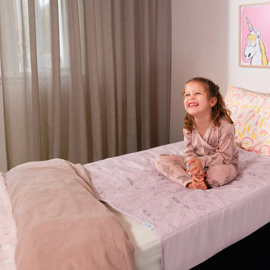 Child sitting on a bed with a unicorn-themed pillow and brolly sheet in a bedroom setting