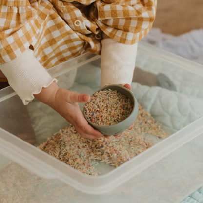 Child engaging in hands-on sensory play using wooden scoop and rainbow rice in sensory bin – supports emotional regulation and NDIS-eligible therapy goals